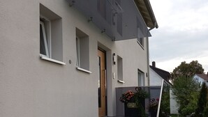 Un bâtiment moderne à l’extérieur gris, avec un escalier en pierre grise menant à une porte, et un petit espace jardin agrémenté d’un banc et d’une table.