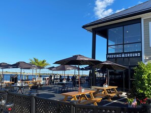 Un espace extérieur aménagé avec des tables et des parasols, une vue sur la mer et un bâtiment portant l’inscription « TERRAIN D’ESSAIS ».