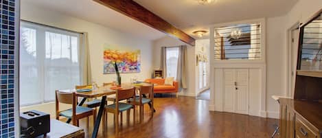 Un comedor amplio con suelo de madera, una mesa y sillas. Hay un ventanal con cortinas, un cuadro en la pared y una escalera que sube a un nivel superior.
