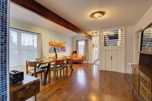 A spacious dining area with wooden flooring, a dining table, and chairs. There is a large window with curtains, a painting on the wall, and a staircase leading to an upper level.
