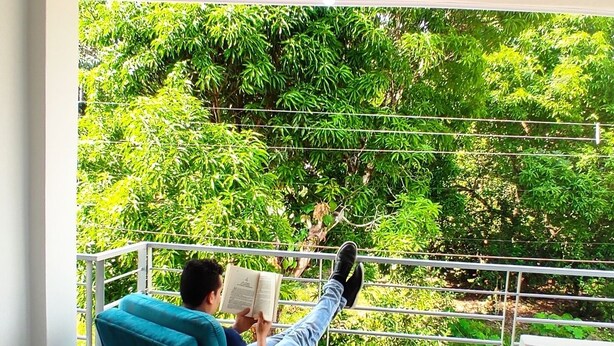 Una persona leyendo un libro en un sofá azul, en una habitación con vistas a abundante vegetación.