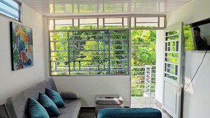 Un salón con un sofá gris, cojines azul verdosos, un pequeño reposapiés, un televisor montado en la pared y una ventana con vistas a zonas verdes.