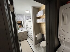 Une petite salle de bain avec des toilettes blanches, une étagère en bois où sont pliées des serviettes, un miroir et un mur à motifs.