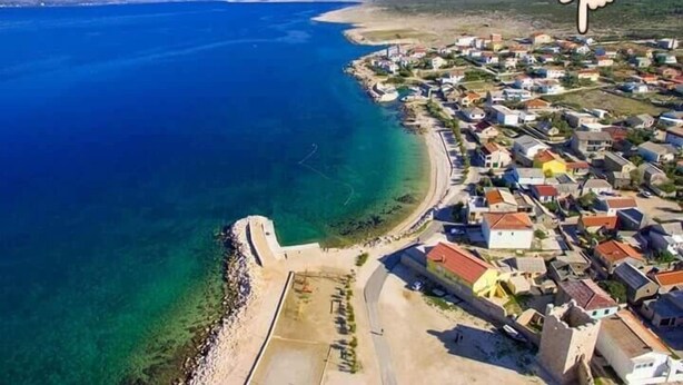 An aerial view of a coastal town with buildings along the shoreline, a clear blue sea, and a mountainous backdrop.