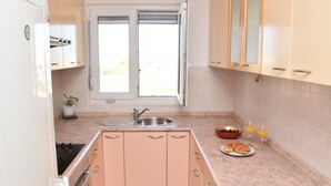 A kitchen with a sink, countertop, and cabinets. There is a window with blinds, a potted plant, and a bowl of fruit and two glasses of orange juice on the counter.