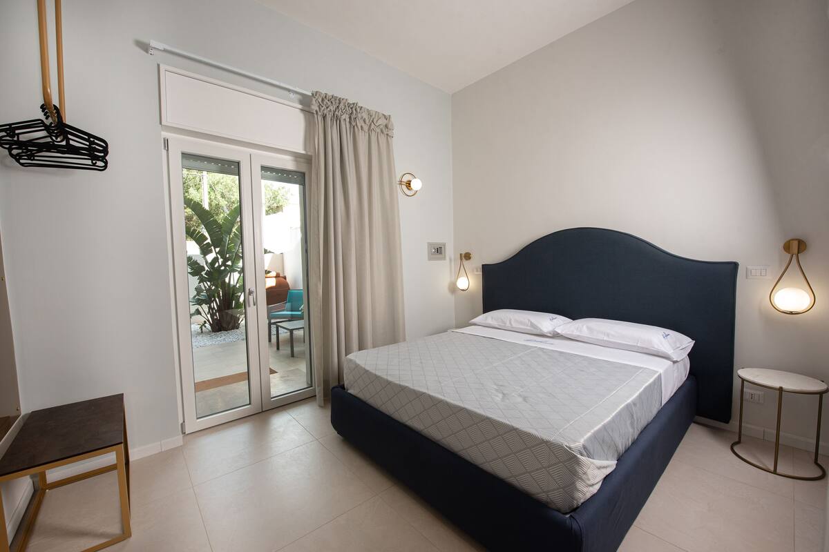 Un dormitorio con cama, una silla, una mesita de noche, una lámpara y un ventanal corredera que da a un espacio exterior.