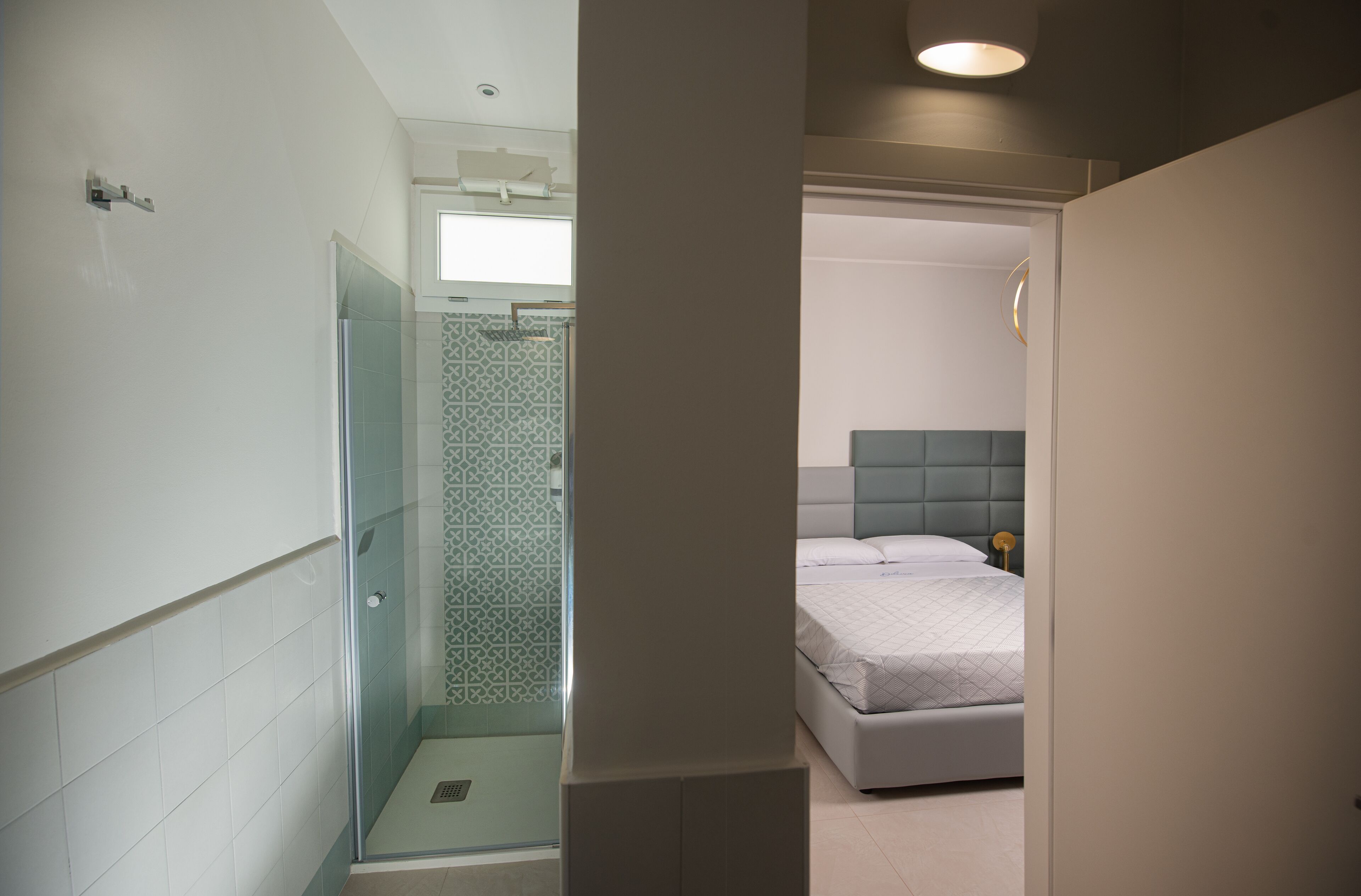 Une salle de bain moderne avec une douche à l’italienne en verre, un mur décoré d’un motif et un lit avec une tête de lit dans la pièce adjacente.