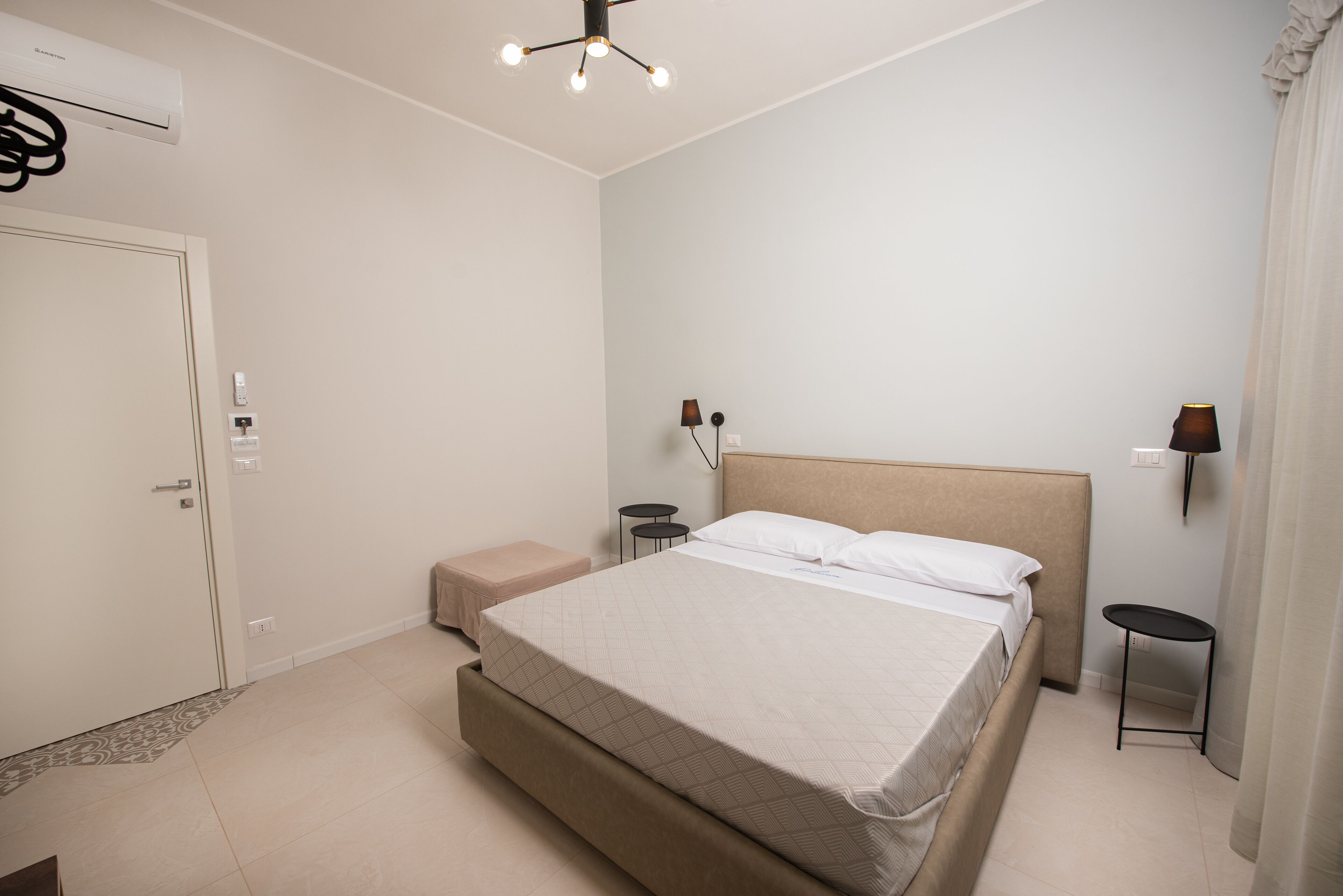 Un dormitorio con una cama, mesitas de noche, una lámpara de pared, aire acondicionado y una puerta.