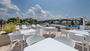 Una terraza en la azotea con mesas y sillas blancas, un banco de madera con cojines azules y una zona de piscina.