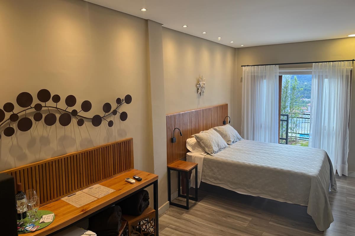 Quarto de hotel com cama, escrivaninha de madeira, objeto decorativo na parede e vista para o exterior.