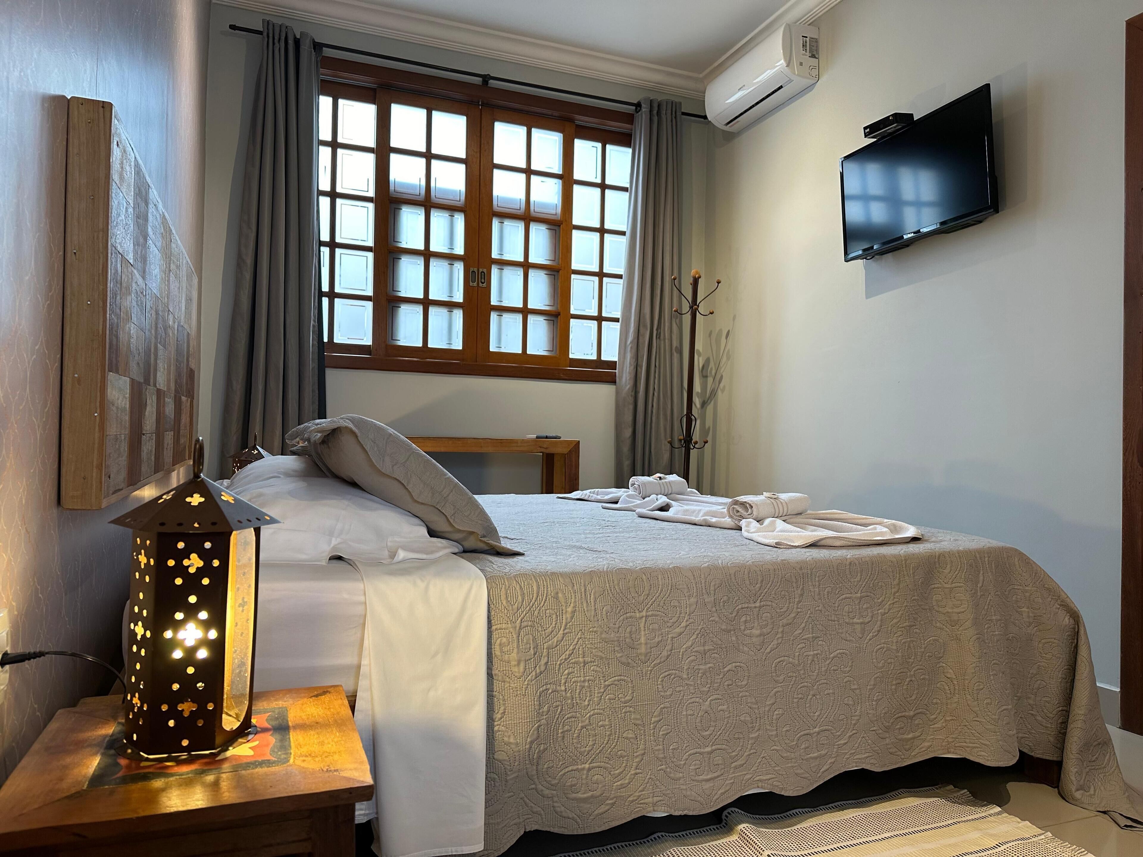 A neatly made bed with a patterned bedspread, a bedside table with a decorative lantern, a flat-screen TV mounted on the wall, and a window with curtains.