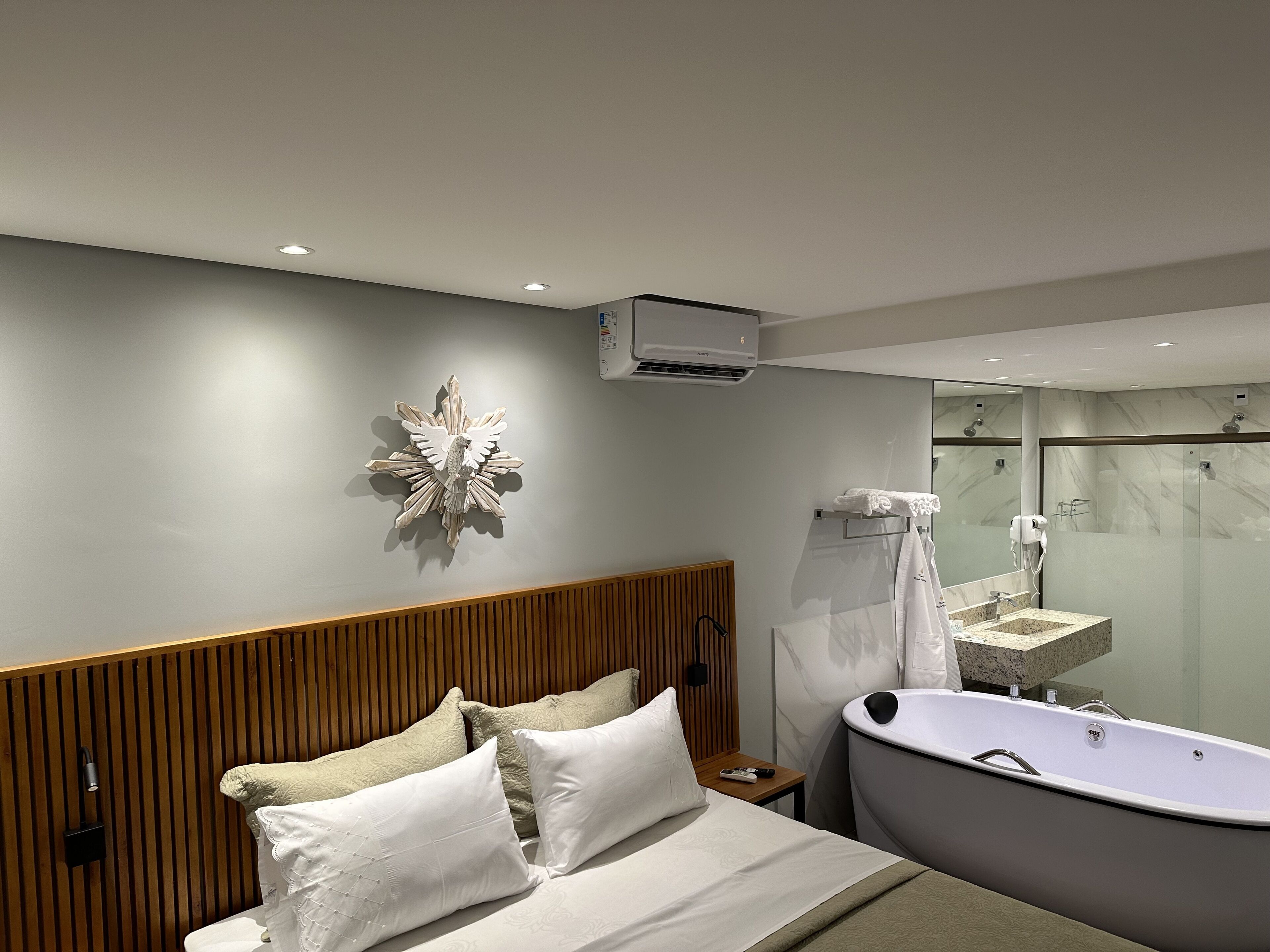 Una habitación de hotel moderna con una cama grande, un cabecero de madera, aire acondicionado empotrado, un tocador con espejo y zona de ducha independiente.