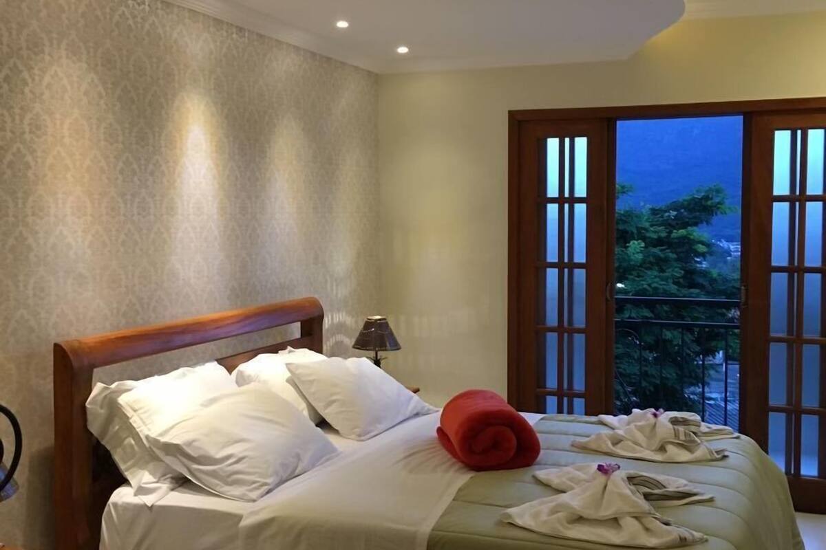 Quarto de hotel com cama, mesa de cabeceira, luminária e vista do exterior através de uma grande janela.