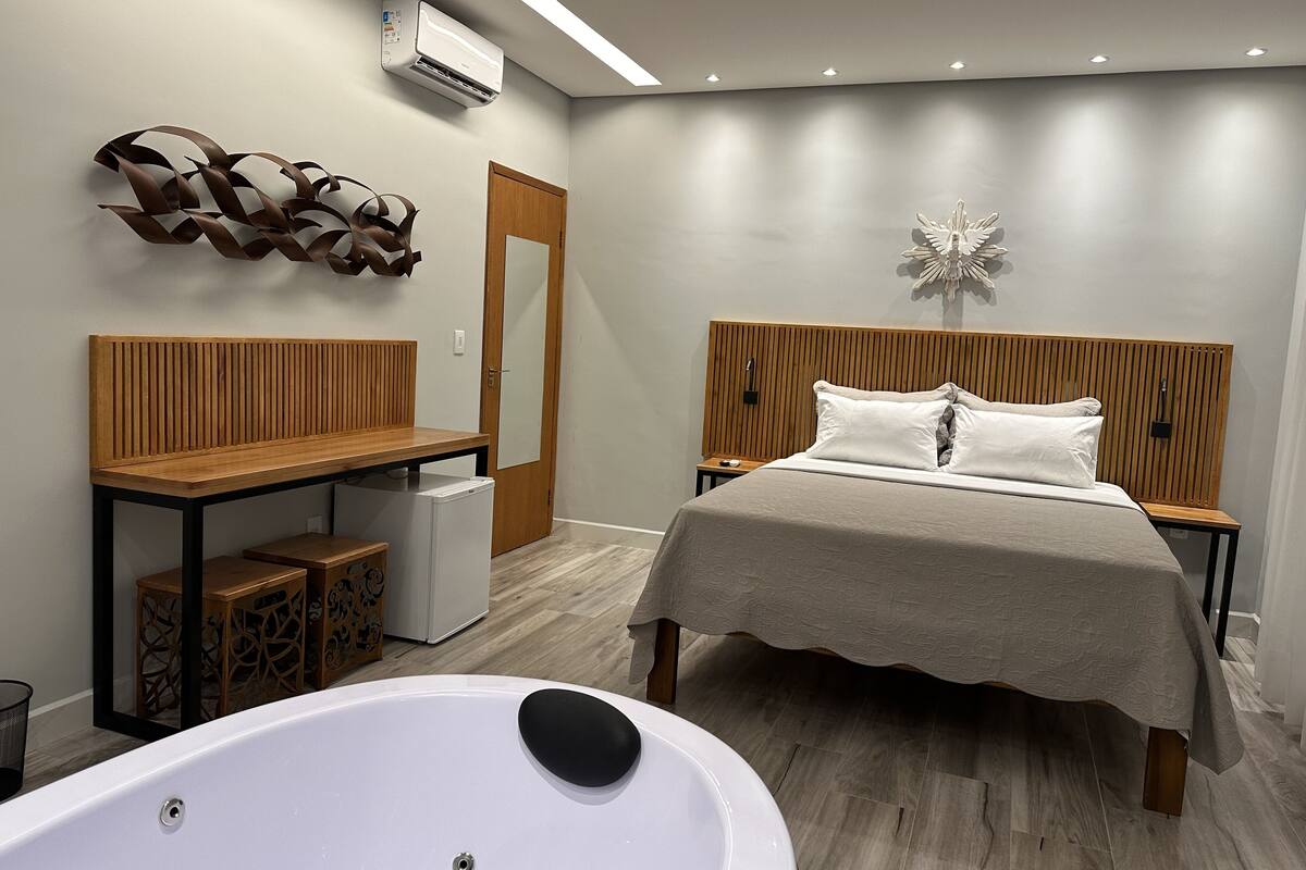 Quarto moderno com cama de madeira, um banco, uma mesinha, uma banheira branca e um ar condicionado fixado na parede.