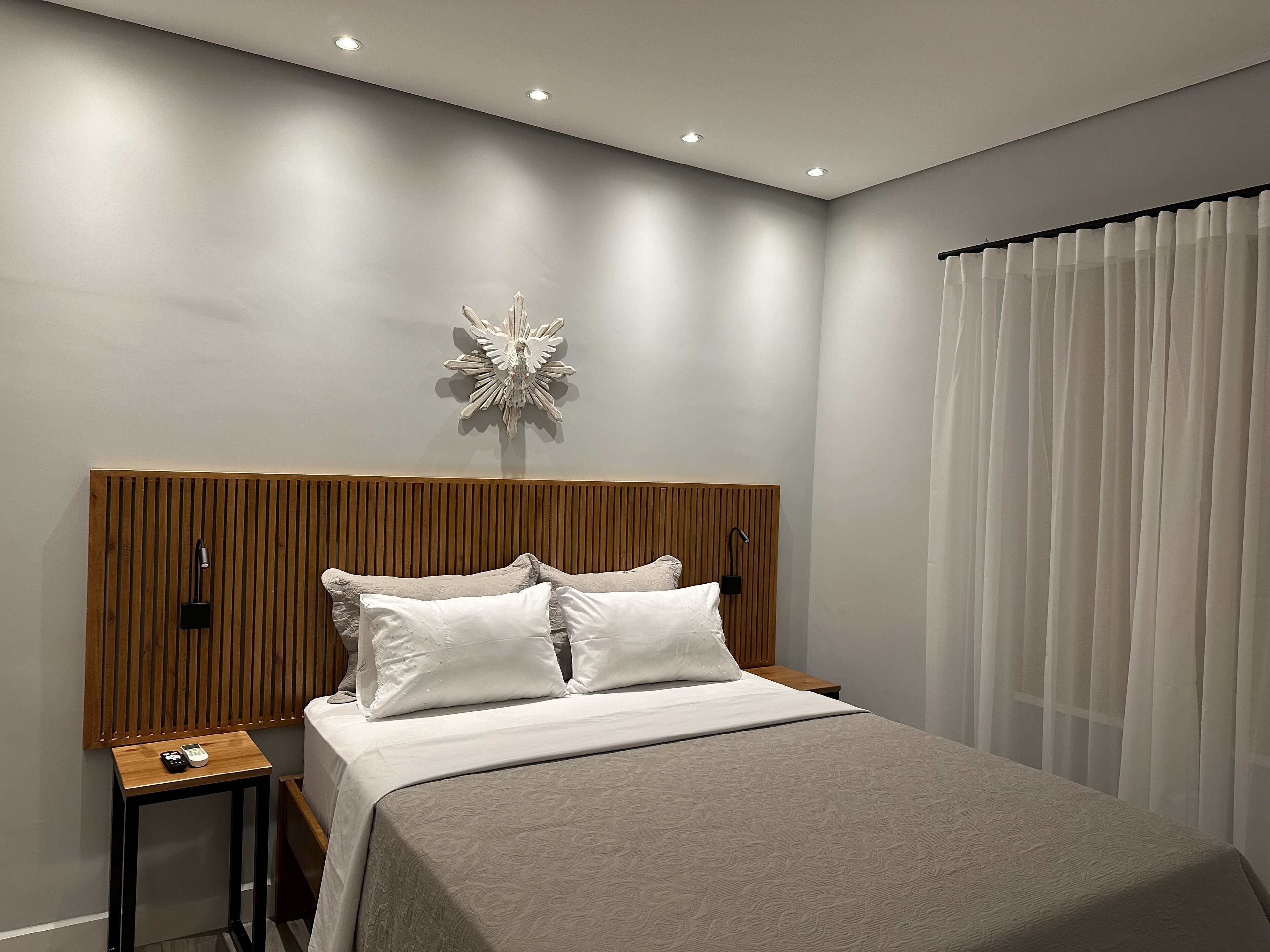 Une chambre à coucher moderne avec une tête de lit en bois, un grand lit avec une literie blanche, une table de chevet avec une télécommande et un élément décoratif mural.