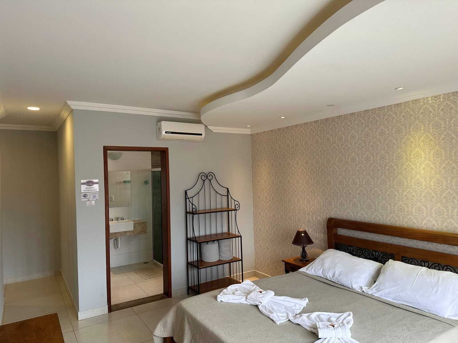 A hotel room with a bed, a nightstand, a lamp, a shelf with towels, and an air conditioning unit.