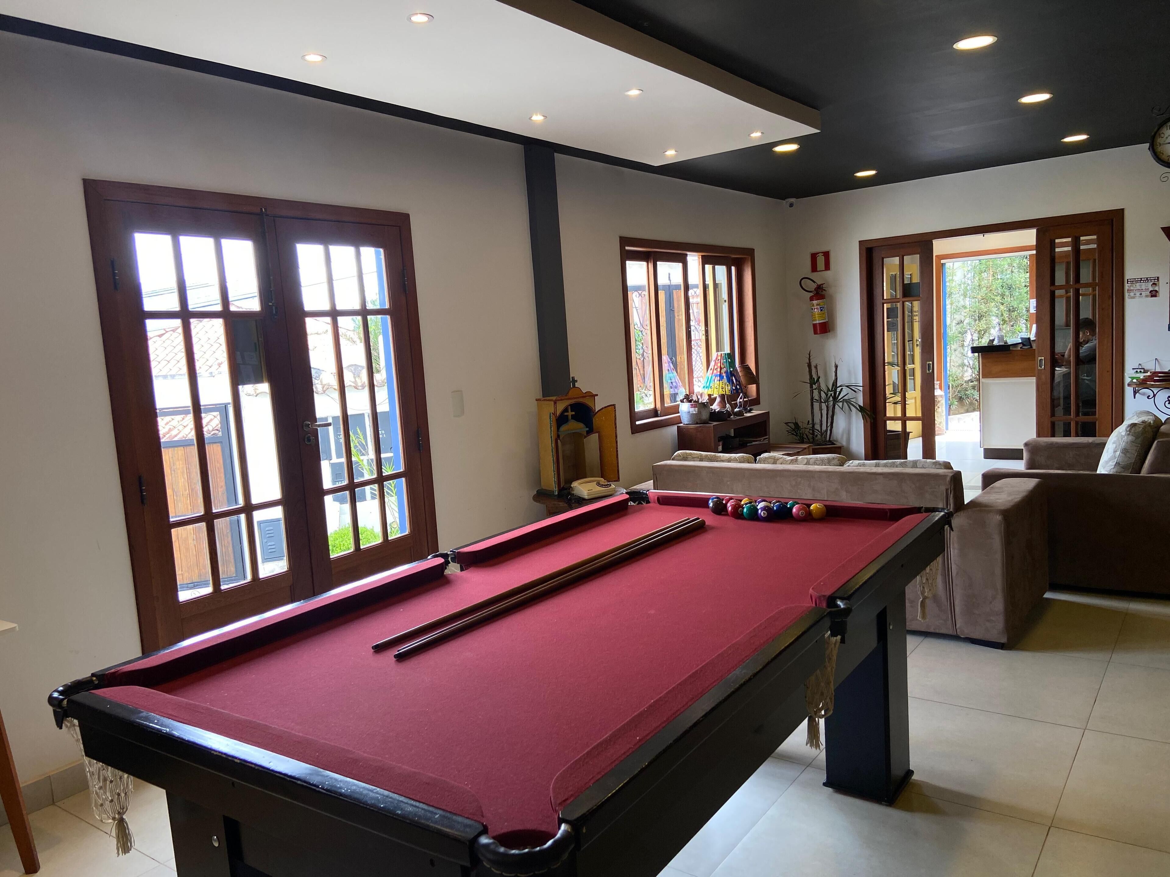 Una mesa de billar con superficie de fieltro rojo, un mueble de madera con una cruz, y una ventana con vistas a follaje.