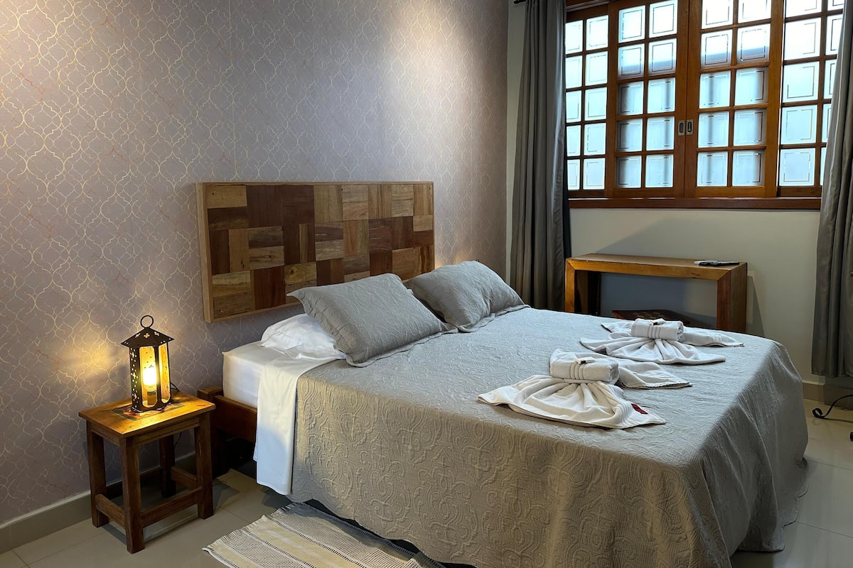 Cama bem arrumada com travesseiros, mesa de cabeceira, abajur e janela com cortinas.