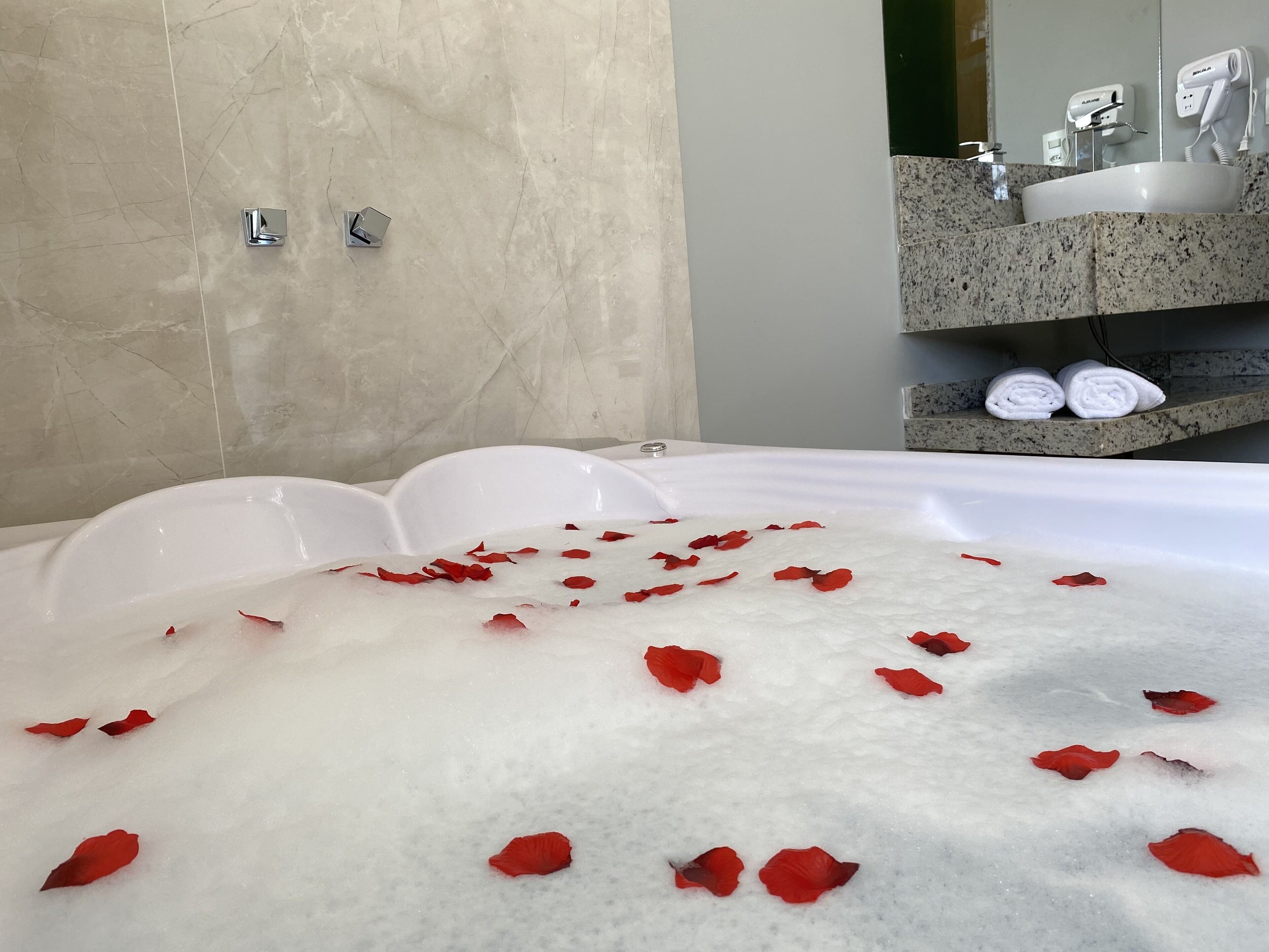 A bathtub filled with bubbles and rose petals, a marble wall, and a bathroom counter with towels and a hairdryer.