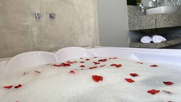 A bathtub filled with bubbles and rose petals, a marble wall, and a bathroom counter with towels and a hairdryer.