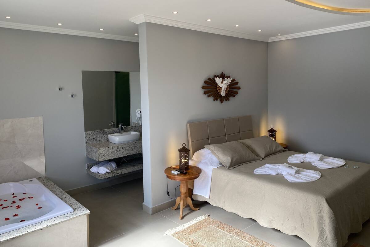 Quarto moderno com uma cama grande, mesa de cabeceira com luminária, banheiro com pia e espelho, e um objeto decorativo na parede.