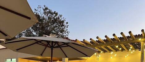 Un espace extérieur aménagé en terrasse, avec un sol en pierre, des chaises en métal blanc et une pergola décorée de guirlandes lumineuses.