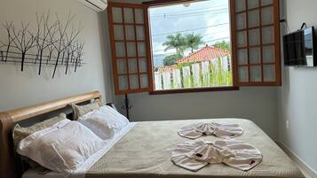 A bedroom with a bed, a window with open shutters, a wall-mounted air conditioner, and a TV on the wall.