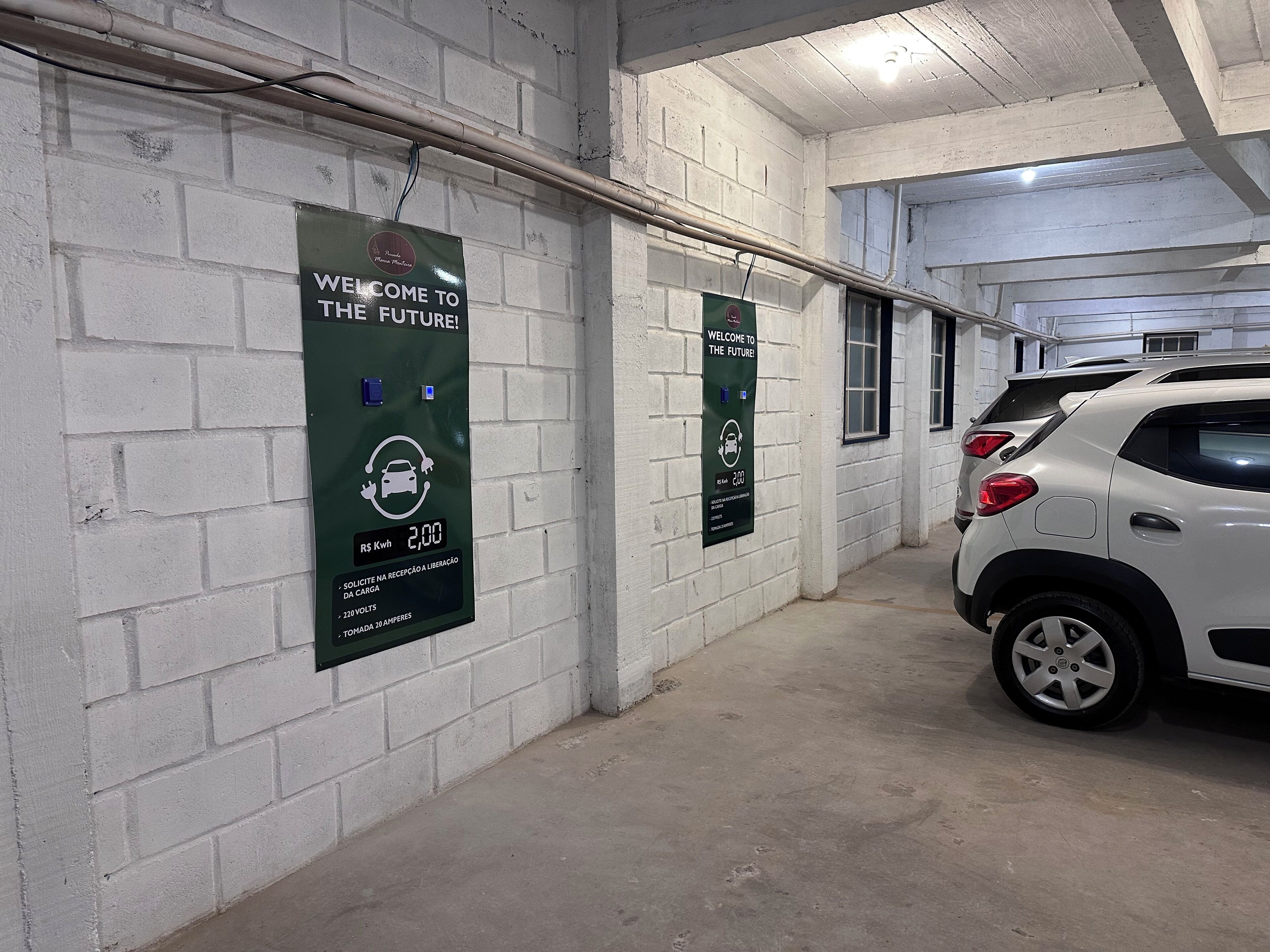 Uma garagem de estacionamento com paredes de tijolos brancos e um carro branco estacionado no interior. Há duas placas na parede que dizem 'Bem-vindo ao Futuro' e exibem um ícone de estação de carregamento com o preço de $2,00 por kWh.