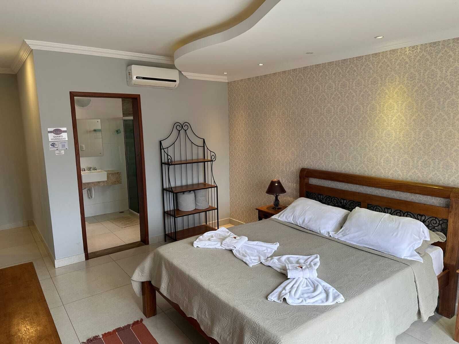 Quarto de hotel com cama, mesa de cabeceira, abajur, guarda-roupa e ar condicionado.
