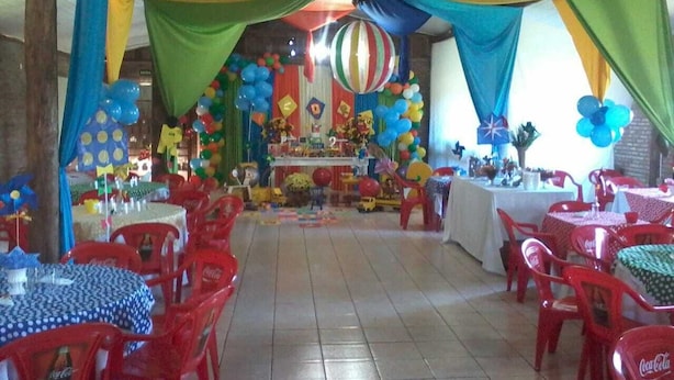 Una habitación con sillas y mesas de plástico rojo, cortinas coloridas y un adorno con forma de globo a rayas.