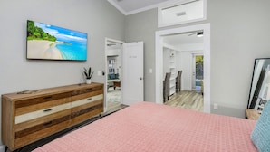 A bedroom with a bed, a TV on the wall, a wooden dresser, and a door leading to another room.