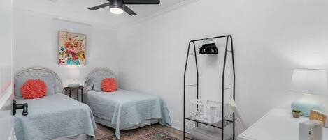 Un dormitorio con dos camas, un ventilador de techo, una mesita de noche con lámpara, una alfombra colorida y un escritorio blanco con lámpara.