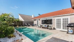 A swimming pool with a tiled deck, outdoor furniture, and a barbecue area.