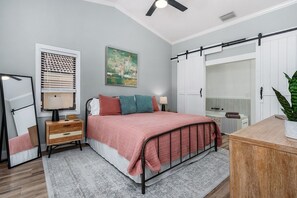 Une chambre à coucher comprenant un lit, une table de chevet, une lampe, un miroir et une grande fenêtre avec des stores.