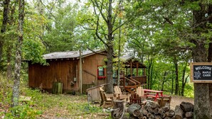 A rustic cabin with a wooden deck, surrounded by dense forest. A signboard reads "Welcome Maple Cabin."