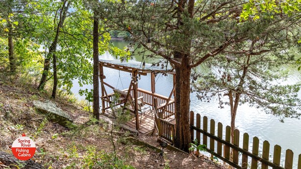 A wooden deck with a railing overlooking a lake, surrounded by trees and a sign indicating 'Spring River Fishing Cabins'.