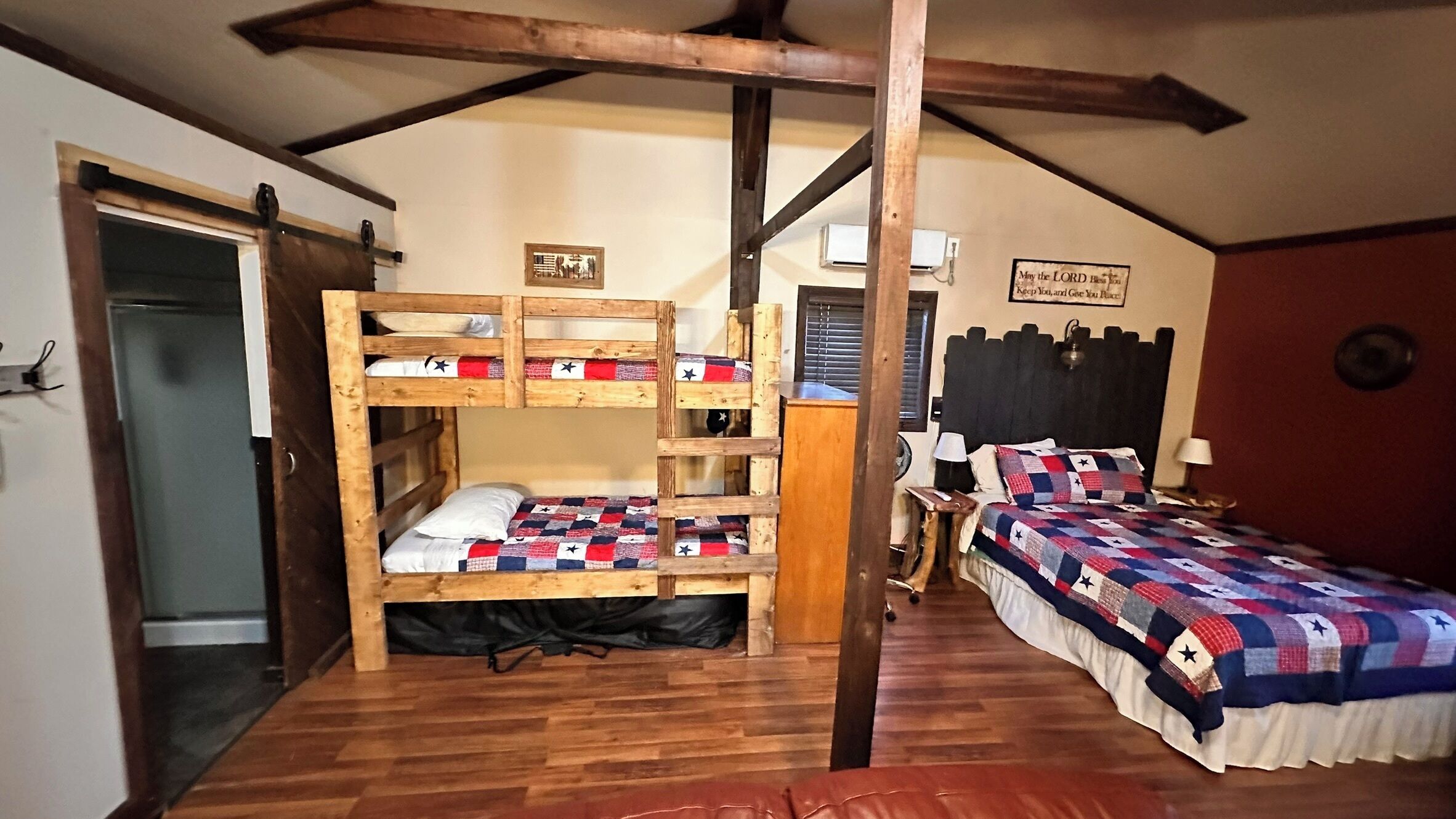 Una habitación con literas, una cama individual, vigas de madera, una puerta, un reloj de pared y un cartel.