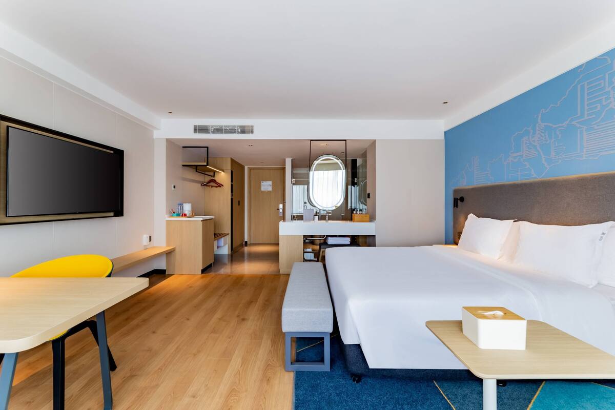 A modern hotel room with a large bed, a desk, a bench, a TV, a bathroom, and a map on the wall.