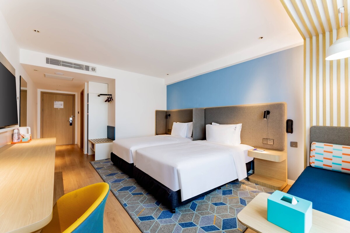 A hotel room with a large bed, a desk, a chair, and a bedside table with a lamp.