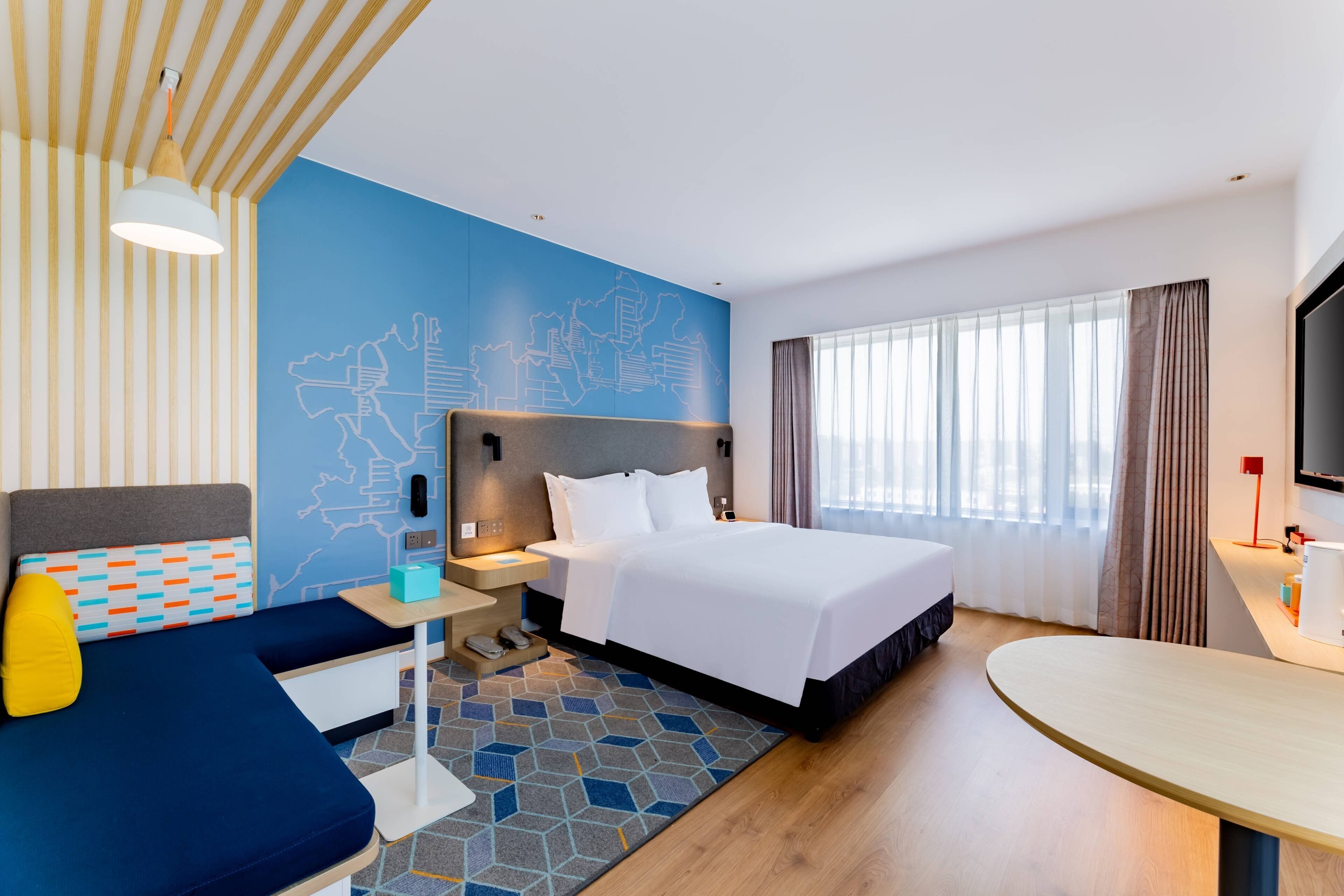 A hotel room with a bed, a desk, a chair, a sofa, a TV, and a window with curtains.