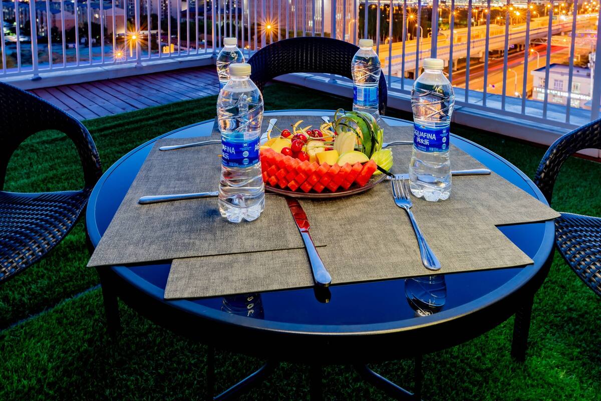 A round table with a fruit platter, water bottles, and cutlery on a terrace with a view of a cityscape at dusk.