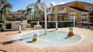 A water play area with a fountain, surrounded by a brick walkway and a fence.