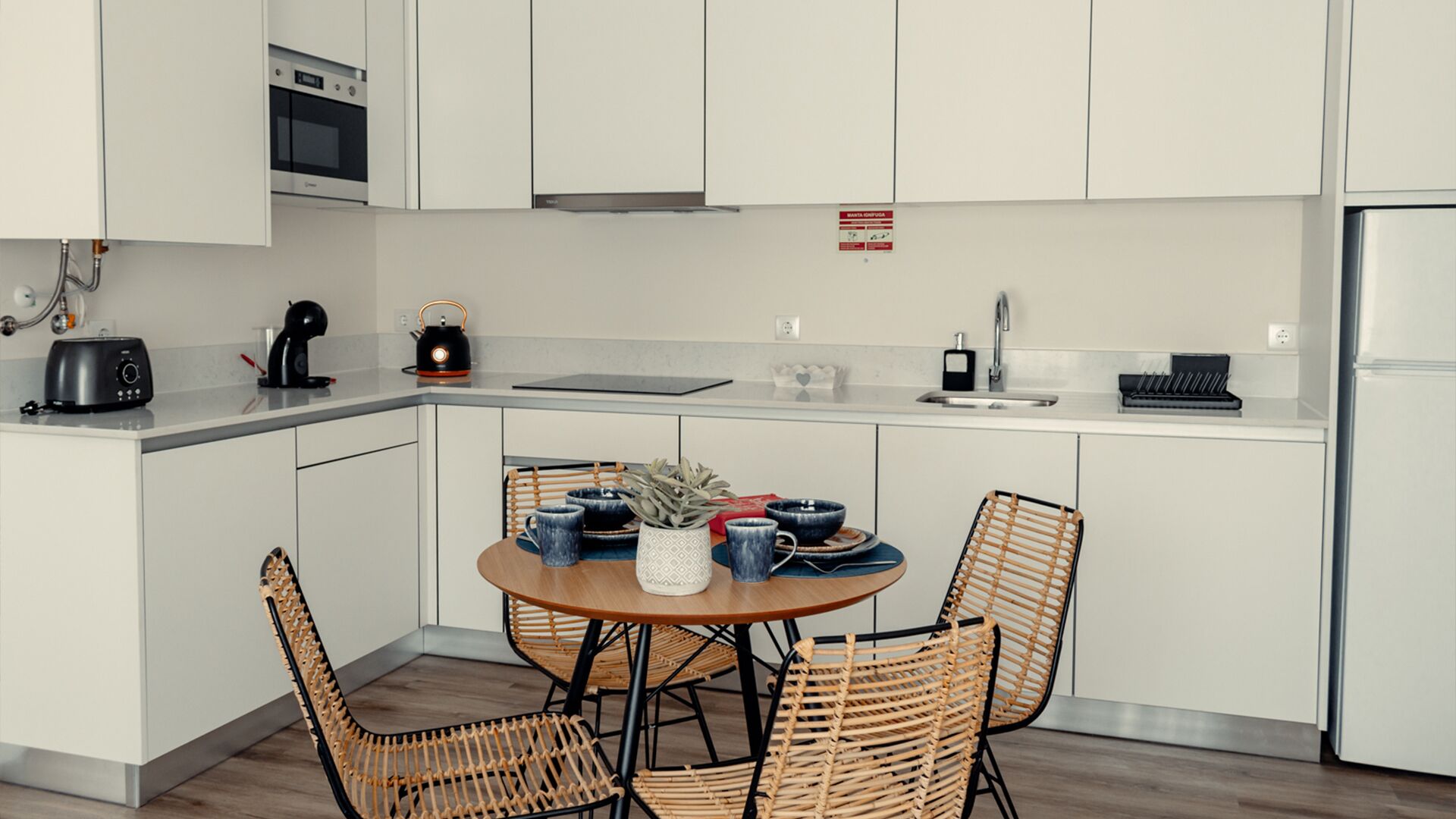 Een moderne keuken met witte kastjes, een inbouwoven, een magnetron, een waterkoker en een spoelbak. Er staat een rond tafel met stoelen en een kleine plant in een pot.