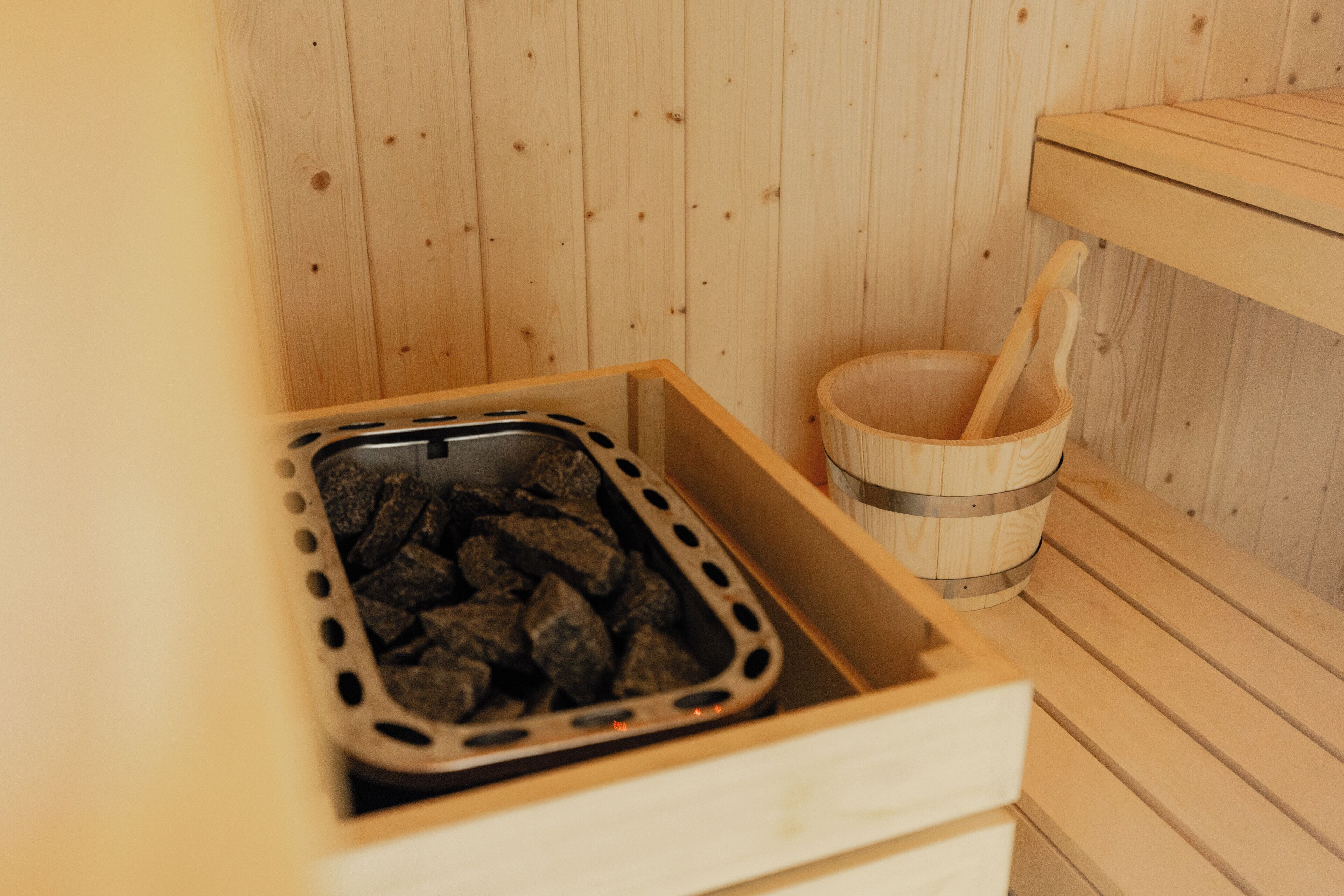 Uma sauna com interior em madeira, um compartimento para carvão e um balde de madeira com uma concha.