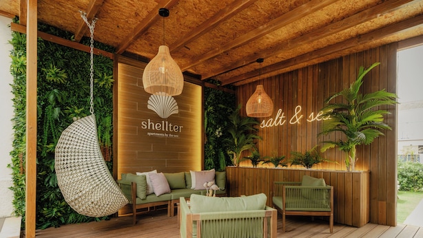 Um deck de madeira com cadeiras penduradas, um sofá e uma parede com vegetação e uma placa com a inscrição 'shellter apartments by the sea'.