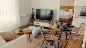 Uma sala de estar moderna com televisão de tela plana, um espaço de refeições com cadeiras de vime e uma cama aconchegante ao fundo.