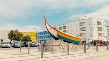 Uma escultura de barco colorido em praça pública, com um muro de pedra e um hotel ao fundo.