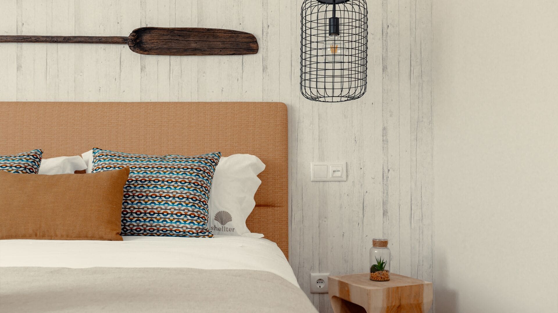 Ein Schlafzimmer mit Holz-Kopfstütze, gemusterten Kissen, einer weißen Bettdecke, einem kleinen Nachttisch mit einem Glasbehälter und einer Wandleuchte.