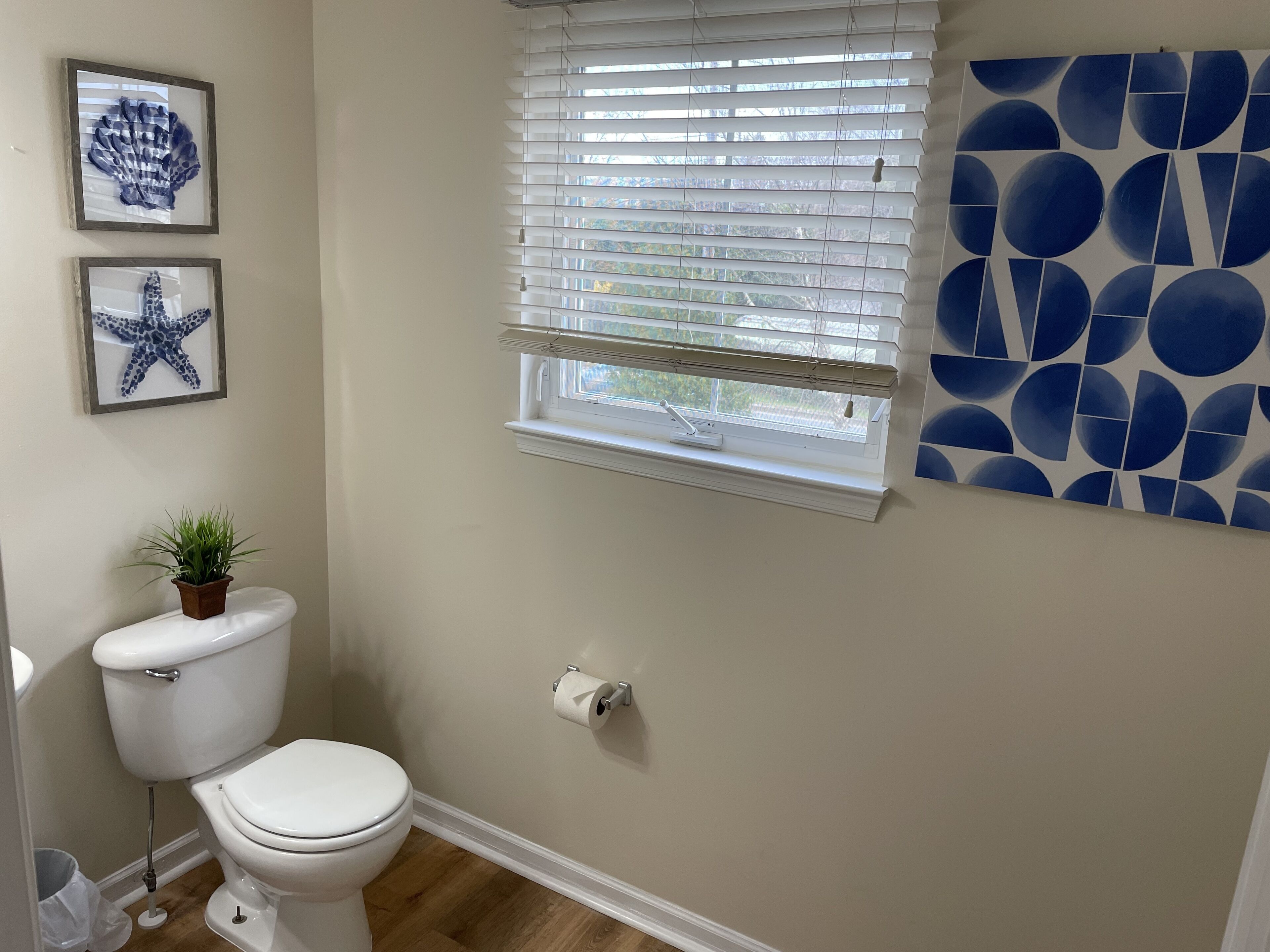 Un baño con inodoro, una ventana con persianas, un dispensador de papel higiénico en la pared y dos cuadros enmarcados.