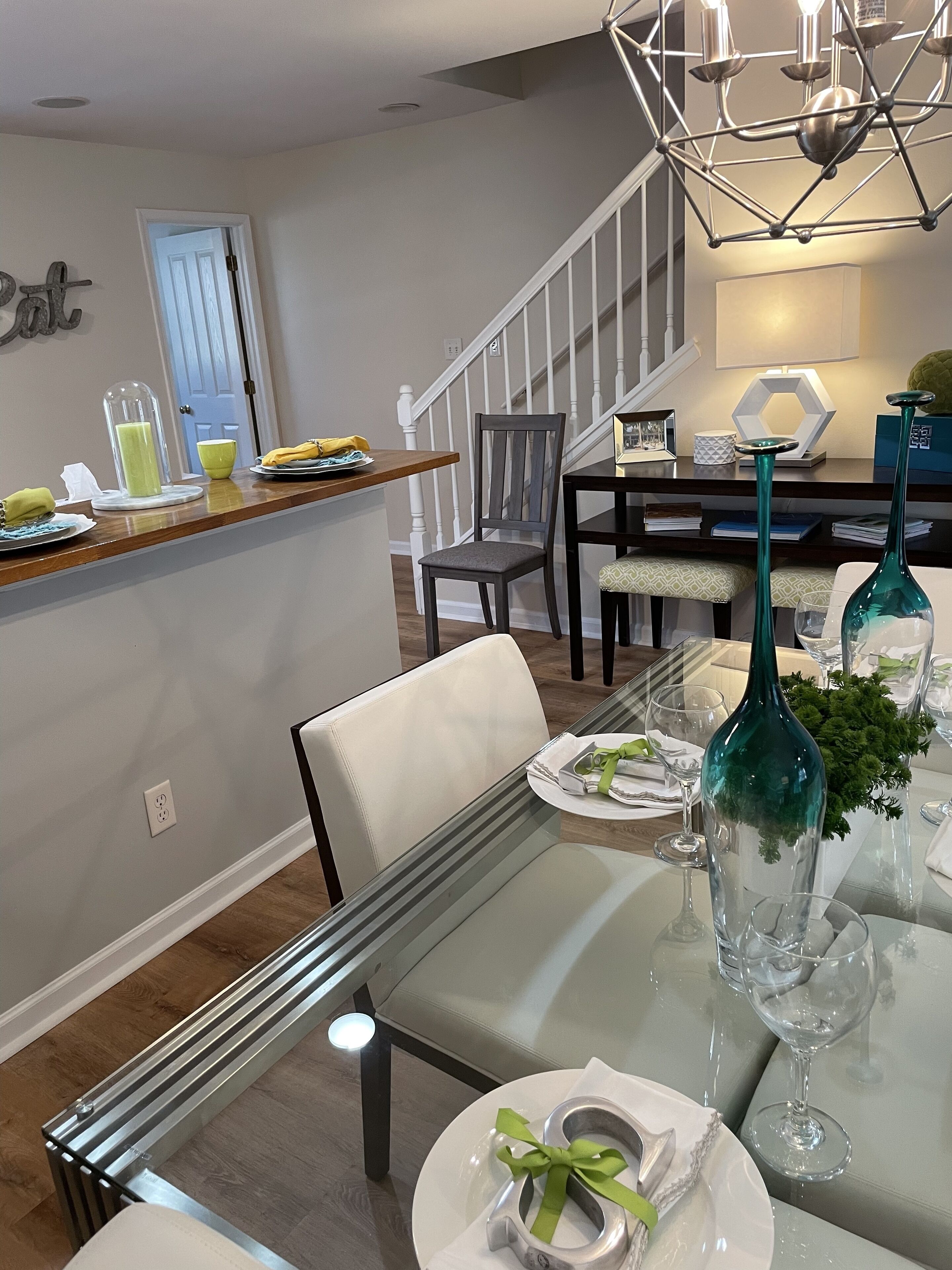 A dining area with a glass table set for two, a chair, a decorative vase, and a staircase in the background.